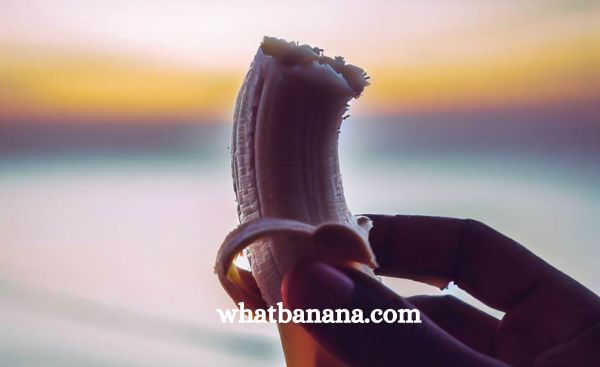 A close-up image of a person's hand holding a peeled banana, with a serene backdrop of a sunset